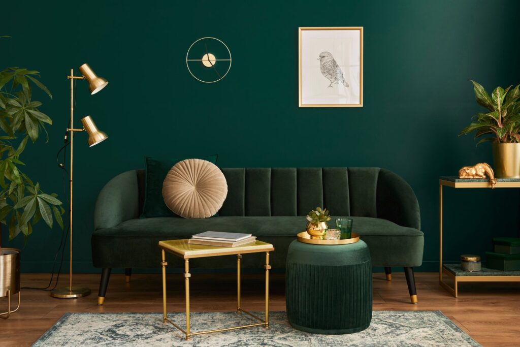 Ein gemütliches Wohnzimmer mit einem grünen Sofa und einer dunkelgrünen Wand, das mit stylischen goldenen Elementen dekoriert wurde. Ein farblich passender Teppich ergänzt das Design.