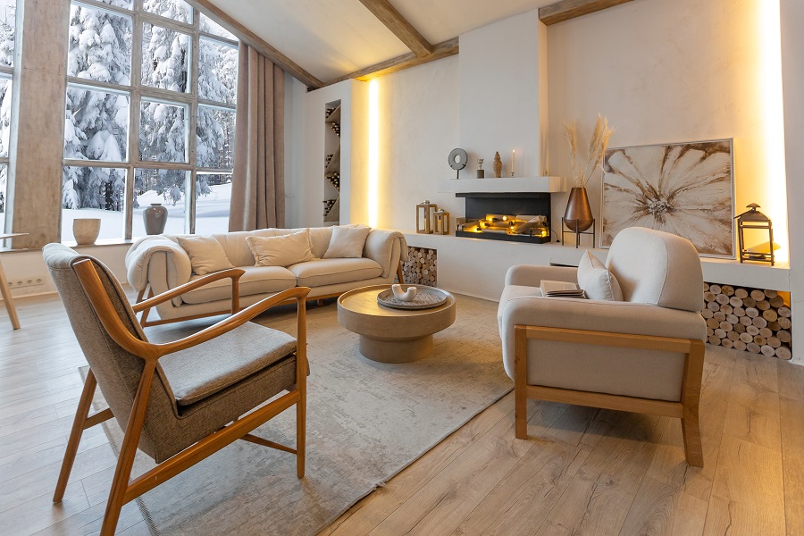 Ein gemütlich eingerichtetes Wohnzimmer in hellen Tönen und mit indirektem Licht. Durch die großen Fenster blickt man auf eine verschneite Winterlandschaft